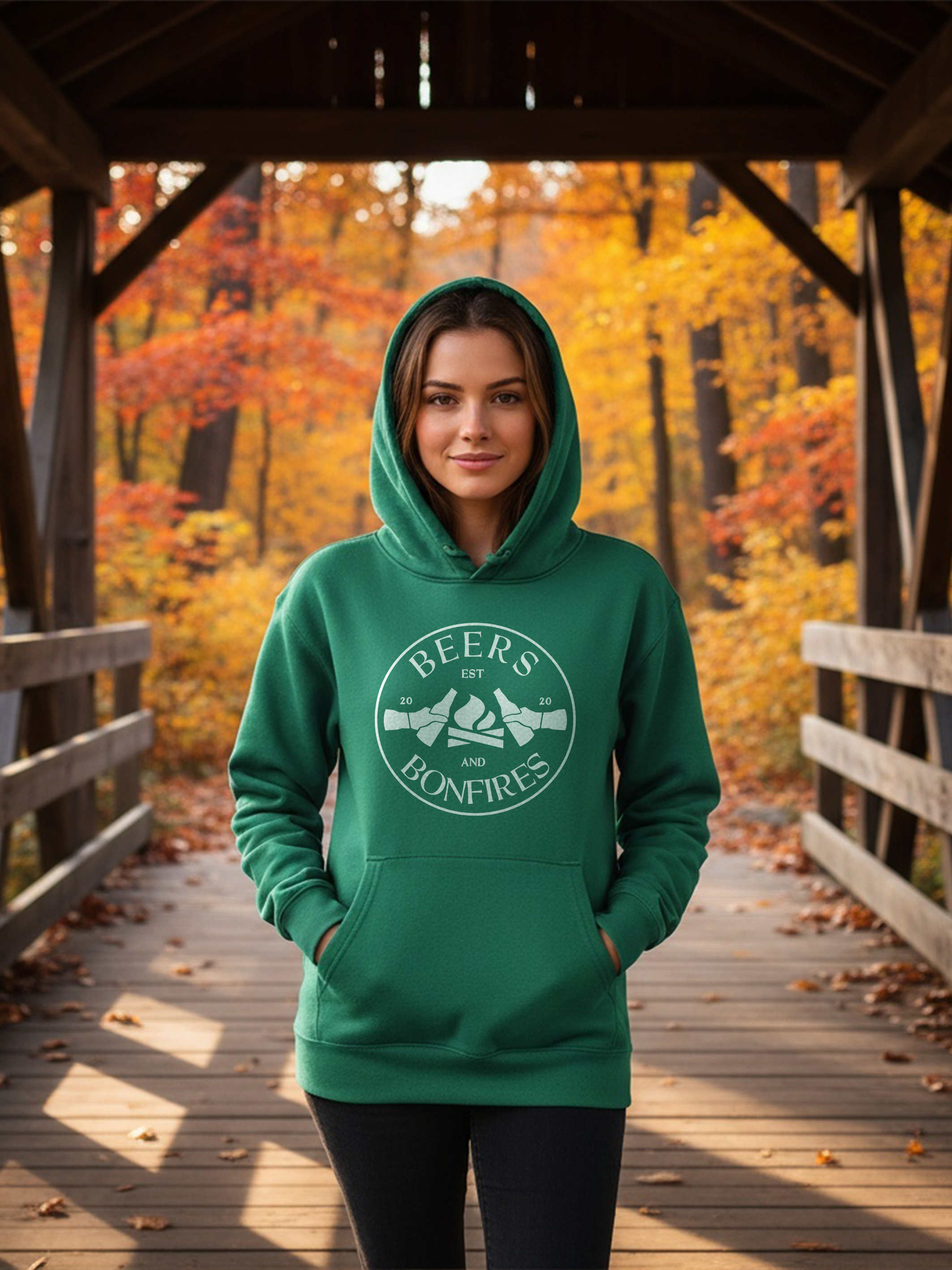 Lady wearing a green hoodie with a logo and text, standing on a wooden bridge with autumn foliage in the background.