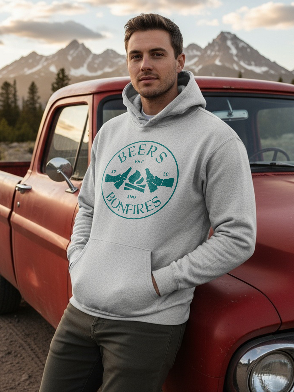 Man wearing a hoodie with Beers and Bonfires design, standing next to a red truck with mountains in the background.